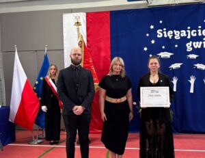 Wręczenie Złotego Indeksu Uczelni Metropolitalnej Trzy osoby stoją podczas szkolnej uroczystości zakończenia roku szkolnego. Uczennica klasy maturalnej stojąca po prawej, trzyma Złoty Indeks Uczelni Metropolitalnej, a w tle widoczne są flagi Polski i Unii Europejskiej oraz dekoracja z widocznym fragmentem napisu "Sięgajcie griazd".