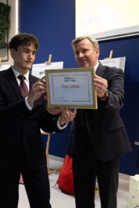 Wręczenie Złotego Indeksu Uczelni Metropolitalnej Mężczyzna i uczeń klasy maturanej, ubrani w garnitury, prezentują oprawiony Złoty Indeks Uczelni Metropolitalnej podczas uroczystości zakończenia roku szkolnego.