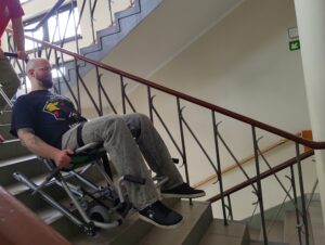 krzesło ewakuacyjne Mężczyzna zjeżdża po schodach na krześle ewakuacyjnym, asekurowany przez drugą osobę. Zdjęcie wykonano wewnątrz budynku, na klatce schodowej. Fotografia ilustruje proces ewakuacji osoby o ograniczonej mobilności przy użyciu specjalistycznego sprzętu.